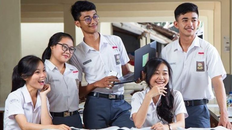 Five students in matching uniforms pose and study together, smiling around a table with notebooks and a laptop in a classroom or library setting.