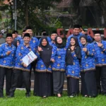 Group of colleagues in matching blue batik uniforms and black caps posing outdoors, giving thumbs up.