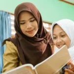 Two young women wearing hijabs sit together at a desk, reading a book in a bright green classroom.