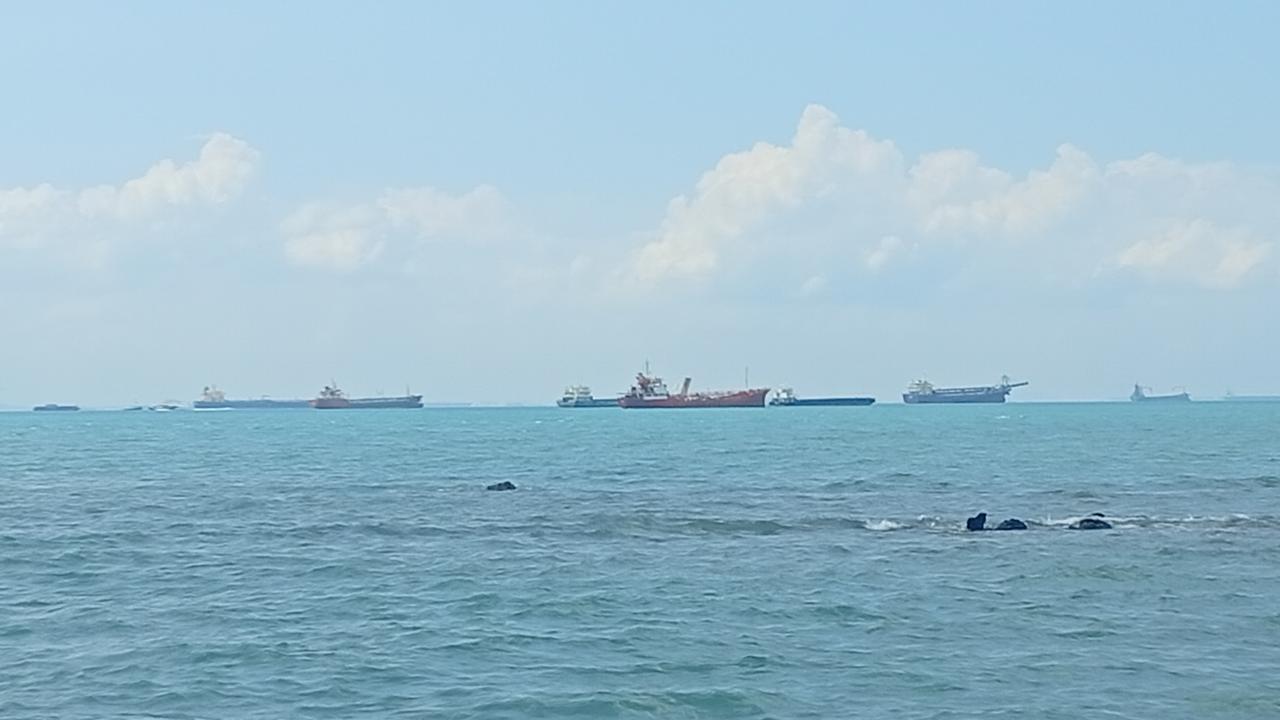 Calm sea with several ships anchored on the horizon beneath a blue sky and scattered clouds.