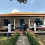 Colonial-style building with orange-tiled roof, white pillars, blue doors, and a veranda; people walk up the stone steps.