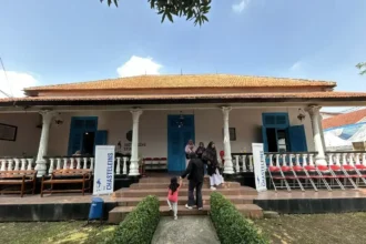 Colonial-style building with orange-tiled roof, white pillars, blue doors, and a veranda; people walk up the stone steps.