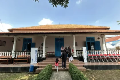 Colonial-style building with orange-tiled roof, white pillars, blue doors, and a veranda; people walk up the stone steps.