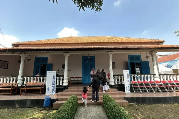 Colonial-style building with orange-tiled roof, white pillars, blue doors, and a veranda; people walk up the stone steps.