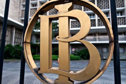 Close-up of a gold circular emblem with an intertwined monogram mounted on a black fence, with a building in the background.