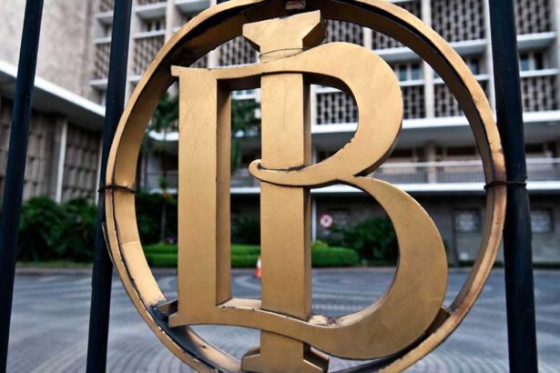 Close-up of a gold circular emblem with an intertwined monogram mounted on a black fence, with a building in the background.