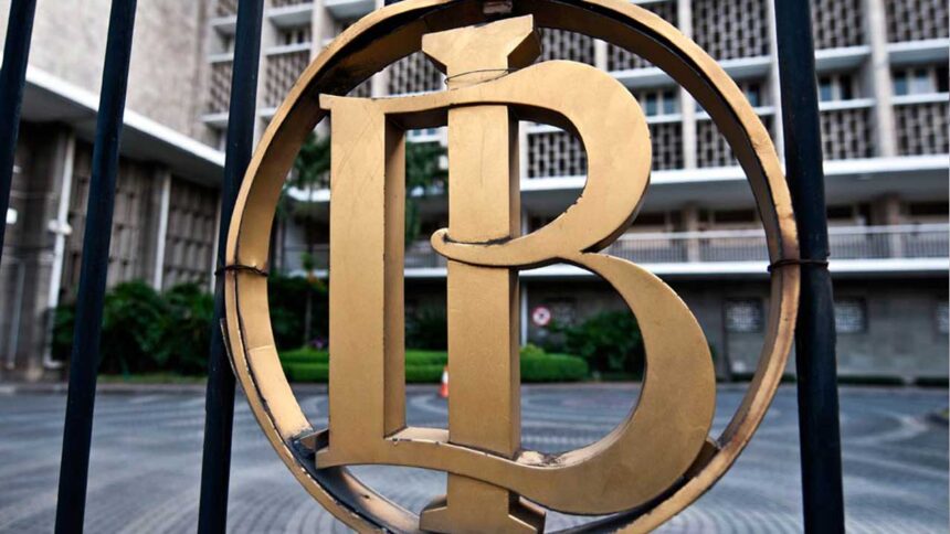 Close-up of a gold circular emblem with an intertwined monogram mounted on a black fence, with a building in the background.