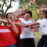 Children in red and white shirts and headbands play tug-of-war outdoors, smiling as they pull a rope together.