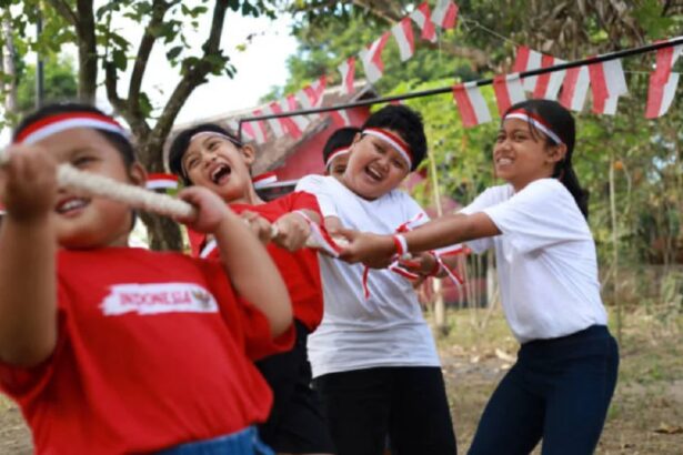Children in red and white shirts and headbands play tug-of-war outdoors, smiling as they pull a rope together.