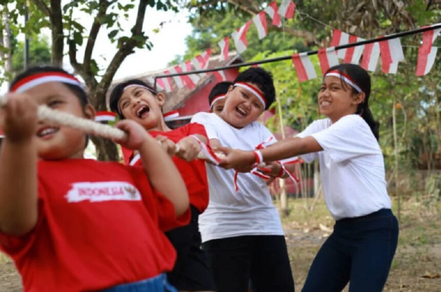 lomba tarik tambang by katadata co id - Mabur.co Children in red and white shirts and headbands play tug-of-war outdoors, smiling as they pull a rope together.