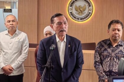 Indonesian official press conference: a man in a dark suit speaks at a podium with the national emblem on the wall.