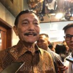 Older man in batik shirt smiles while being surrounded by reporters holding smartphones.