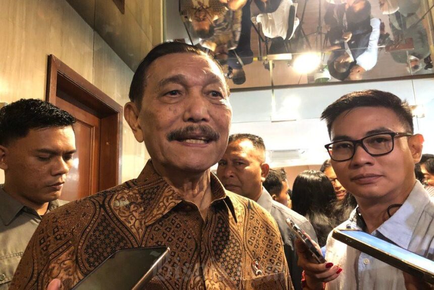 Older man in batik shirt smiles while being surrounded by reporters holding smartphones.