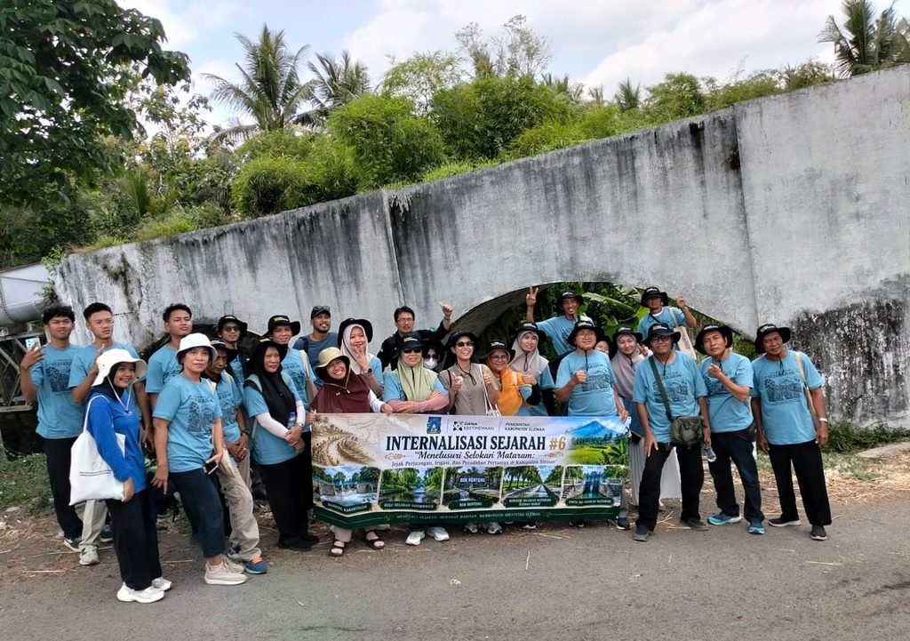 Group of people wearing matching light blue shirts posing outdoors behind a large banner that reads 'Internalisasi Sejarah #6' under a concrete arch with green foliage above.