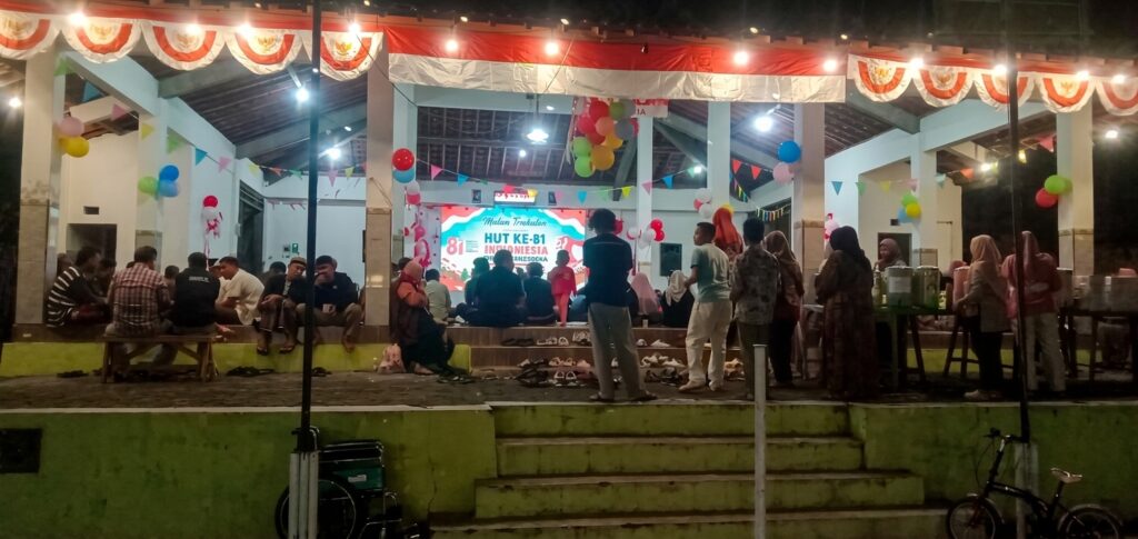 Nighttime community celebration under a decorated pavilion with a stage and banner reading HUT KE-81 Indonesia; people sit and stand around, balloons and bunting, shoes lined up in front.