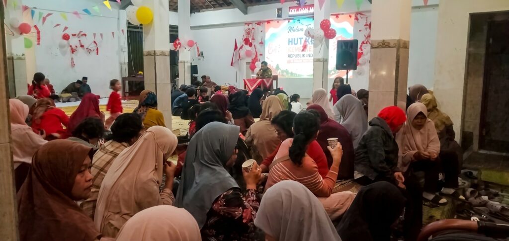 Large indoor community gathering with women in hijabs seated on the floor, a speaker at a podium on a decorated stage in the background.