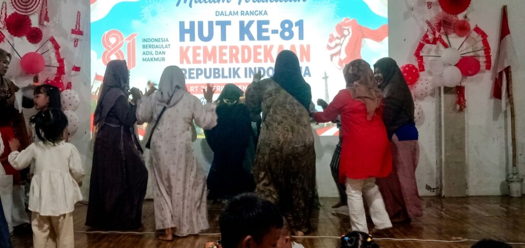 Group of women in hijabs dancing on a stage in front of a banner that reads HUT KE-81 KEMERDEKAAN REPUBLIK INDONESIA, celebrating Independence Day.