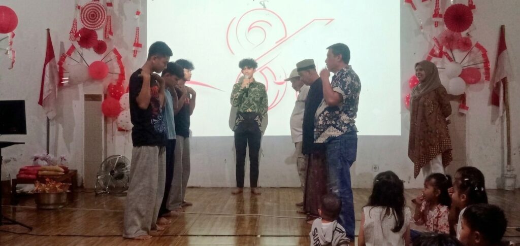 Group of men on stage with one speaking into a microphone at center; a woman in a hijab stands to the right while children sit in front; red-and-white decorations and a projector screen behind.