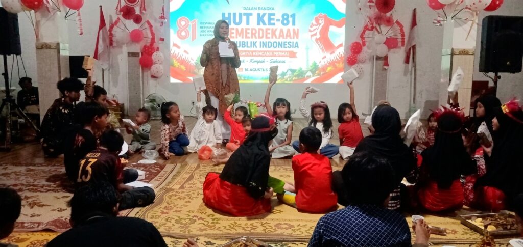 Woman on stage hosting a kids' event with a banner for HUT ke-81 Indonesia; children sit in a circle, holding papers.