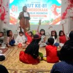 Woman on stage hosting a kids' event with a banner for HUT ke-81 Indonesia; children sit in a circle, holding papers.
