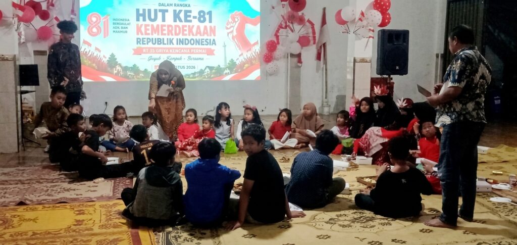 Group of children seated in a circle on a patterned carpet during an Indonesian independence celebration, with balloons and a banner in the background.