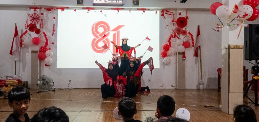 Group of young girls in red and black outfits performing a coordinated dance on a stage, with red-and-white decorations and balloons in the background.