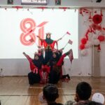 Group of young girls in red and black outfits performing a coordinated dance on a stage, with red-and-white decorations and balloons in the background.