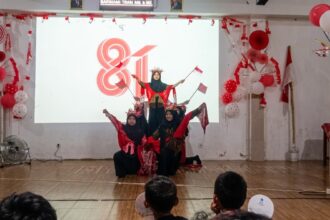Group of young girls in red and black outfits performing a coordinated dance on a stage, with red-and-white decorations and balloons in the background.