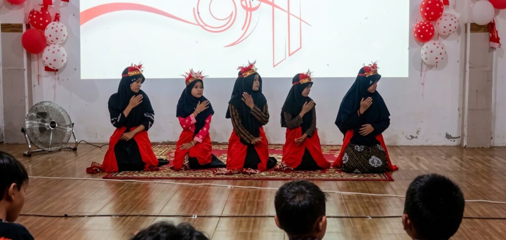 Five girls in black hijabs and red skirts perform a synchronized kneeling dance on a stage, each hand over the chest while facing slightly to the side, on a red patterned rug.