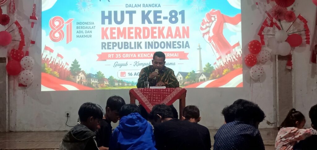 Man in batik shirt speaks at a podium on a decorated stage for Indonesia's 81st Independence Day celebration with a large banner behind him.