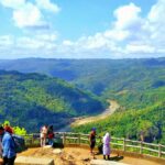 People stand at a wooden railing overlooking a lush valley with a winding river and forested hills under a bright blue sky.