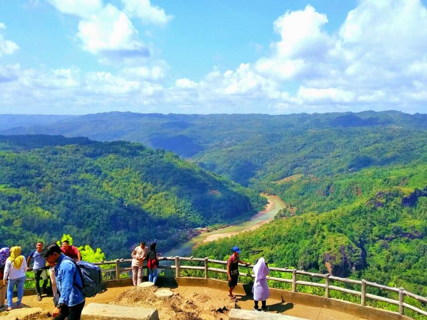 mangunan by travelspromo com - Mabur.co People stand at a wooden railing overlooking a lush valley with a winding river and forested hills under a bright blue sky.