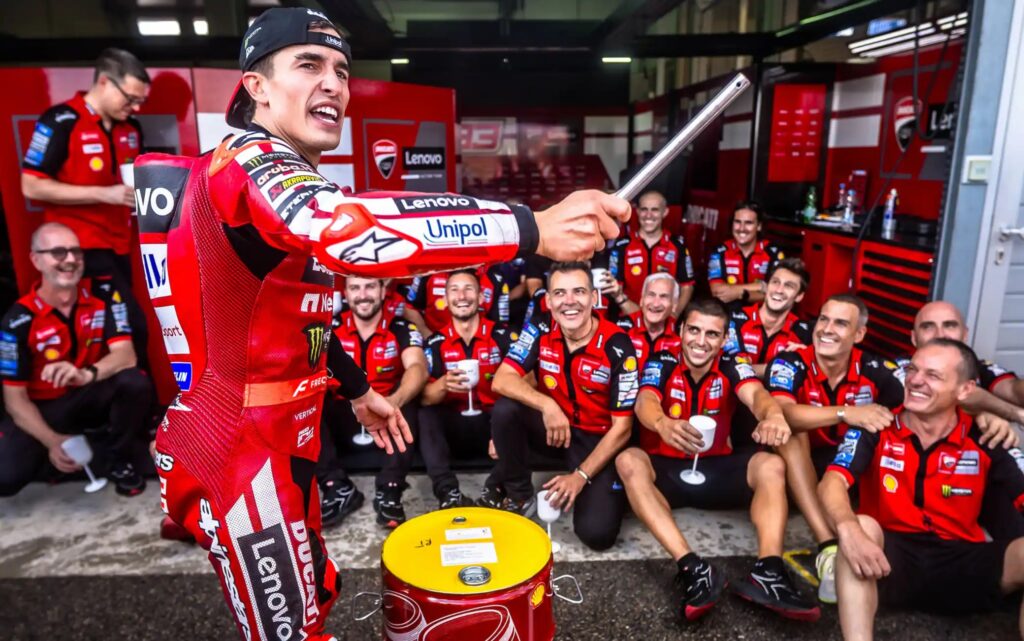 MotoGP team celebration in a red-padded pit garage: a rider in red suit raises a metal bar toward the crowd as teammates sit and stand, smiling and holding cups in the background. Sponsors' logos visible on uniforms.