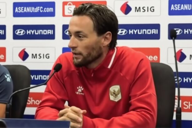 Athlete in a red jacket seated at a press conference, hands clasped, with microphones in front and sponsor logos on the backdrop