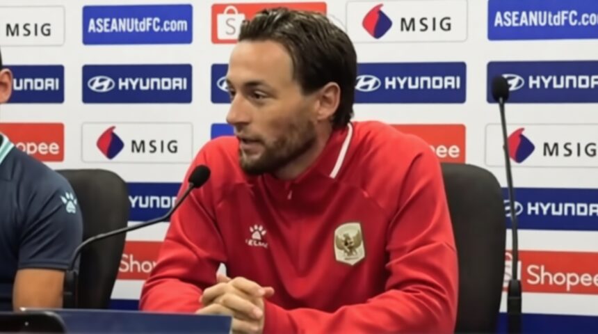 Athlete in a red jacket seated at a press conference, hands clasped, with microphones in front and sponsor logos on the backdrop