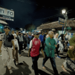Night street scene with a diverse crowd walking together beneath streetlights and shopfronts at night, some wearing colorful clothing.