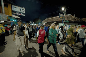 Night street scene with a diverse crowd walking together beneath streetlights and shopfronts at night, some wearing colorful clothing.