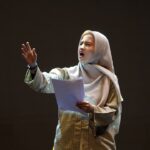Woman wearing a hijab and plaid shirt speaks passionately on a stage, holding papers in front of a dark background.