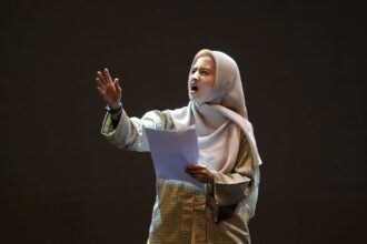 Woman wearing a hijab and plaid shirt speaks passionately on a stage, holding papers in front of a dark background.