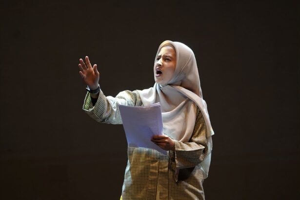 Woman wearing a hijab and plaid shirt speaks passionately on a stage, holding papers in front of a dark background.