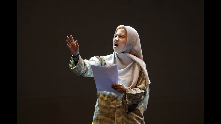 Woman wearing a hijab and plaid shirt speaks passionately on a stage, holding papers in front of a dark background.