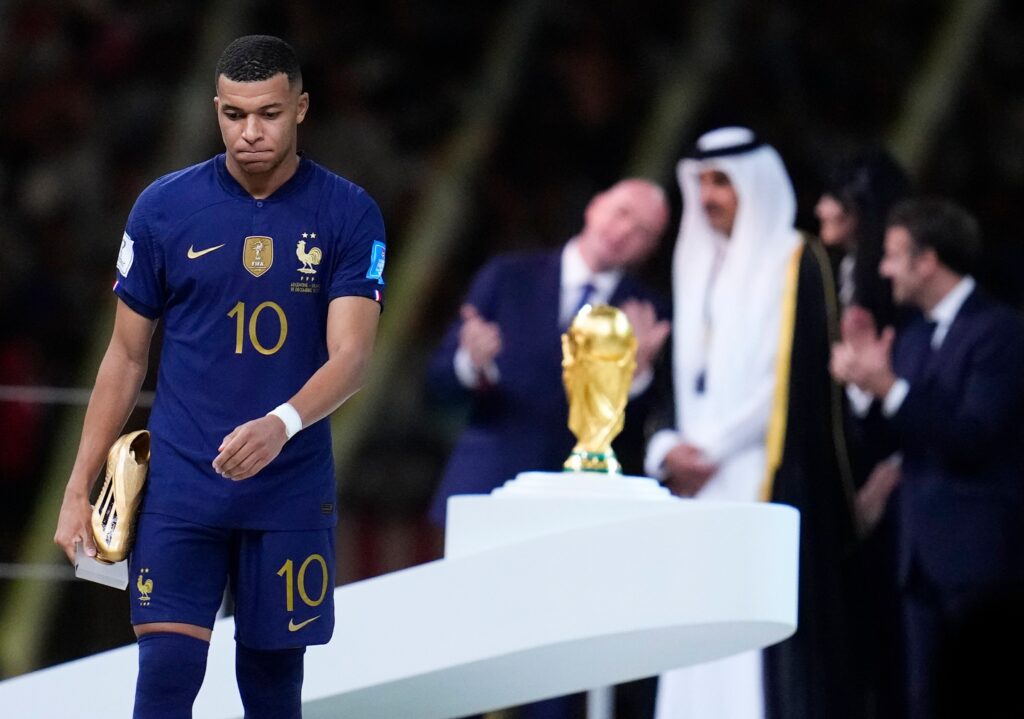 French soccer player in blue kit (number 10) walking past spectators with a gold trophy visible in the foreground.