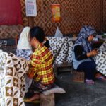 Women sit on low stools weaving patterned fabric in a workshop, with large printed cloths draped over frames nearby.