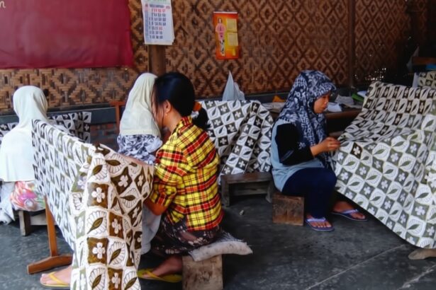 Women sit on low stools weaving patterned fabric in a workshop, with large printed cloths draped over frames nearby.