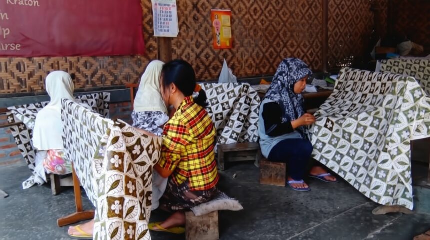 membatik tradisional FIXX - Mabur.co Women sit on low stools weaving patterned fabric in a workshop, with large printed cloths draped over frames nearby.