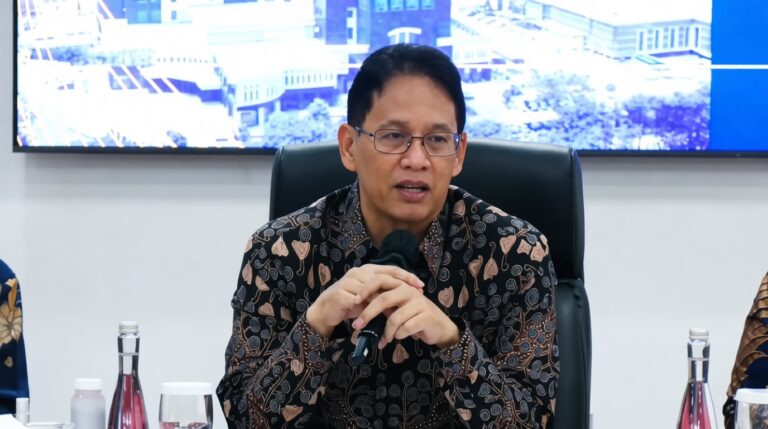 Man in a patterned batik shirt speaks into a handheld microphone at a conference table, with water bottles nearby and a blue display behind him.