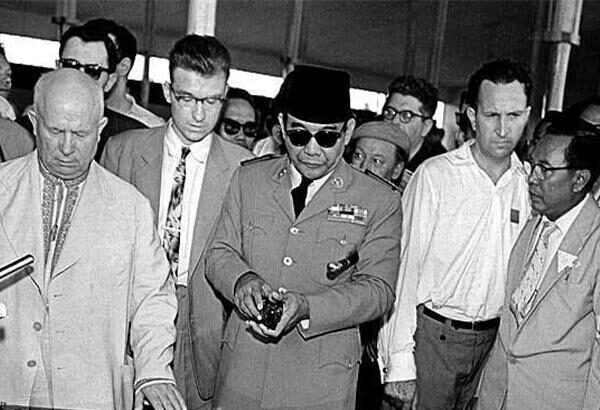 Group of men in suits and a uniformed officer with sunglasses examining a large technical model or control panel at an exhibition, focused on the central display.