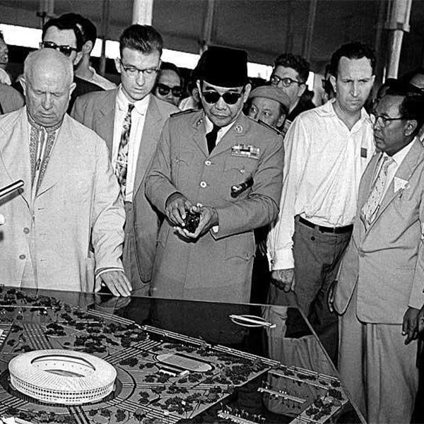 Group of men in suits and a uniformed officer with sunglasses examining a large technical model or control panel at an exhibition, focused on the central display.