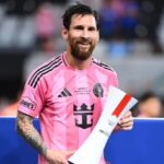 Professional soccer player with a pink jersey and beard, smiling while holding a rolled trophy or certificate at a stadium.