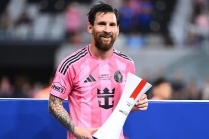 Professional soccer player with a pink jersey and beard, smiling while holding a rolled trophy or certificate at a stadium.